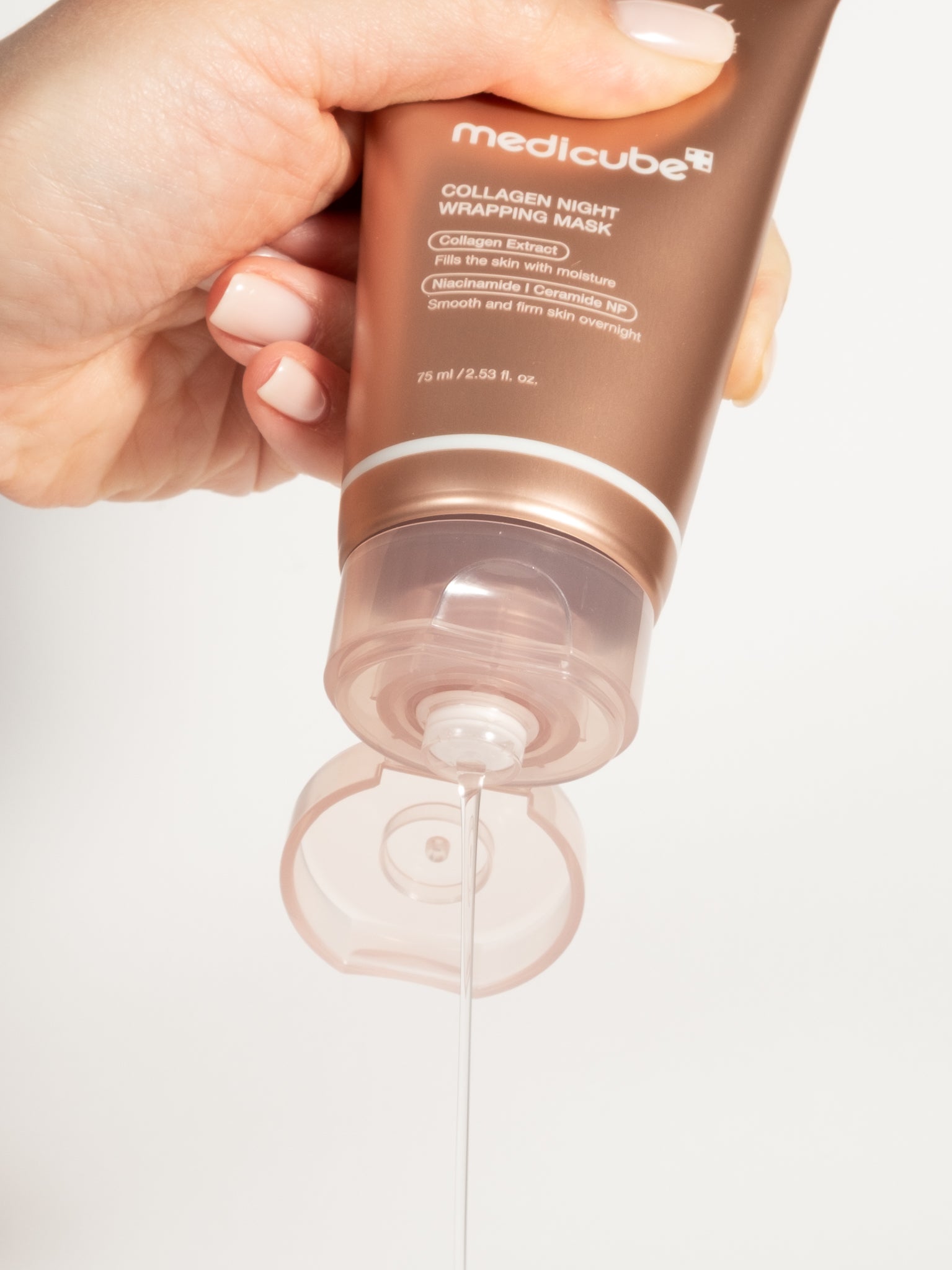 A hand squeezing a clear, gel-like string of Medicube Collagen Night Wrapping Mask from a bronze tube.