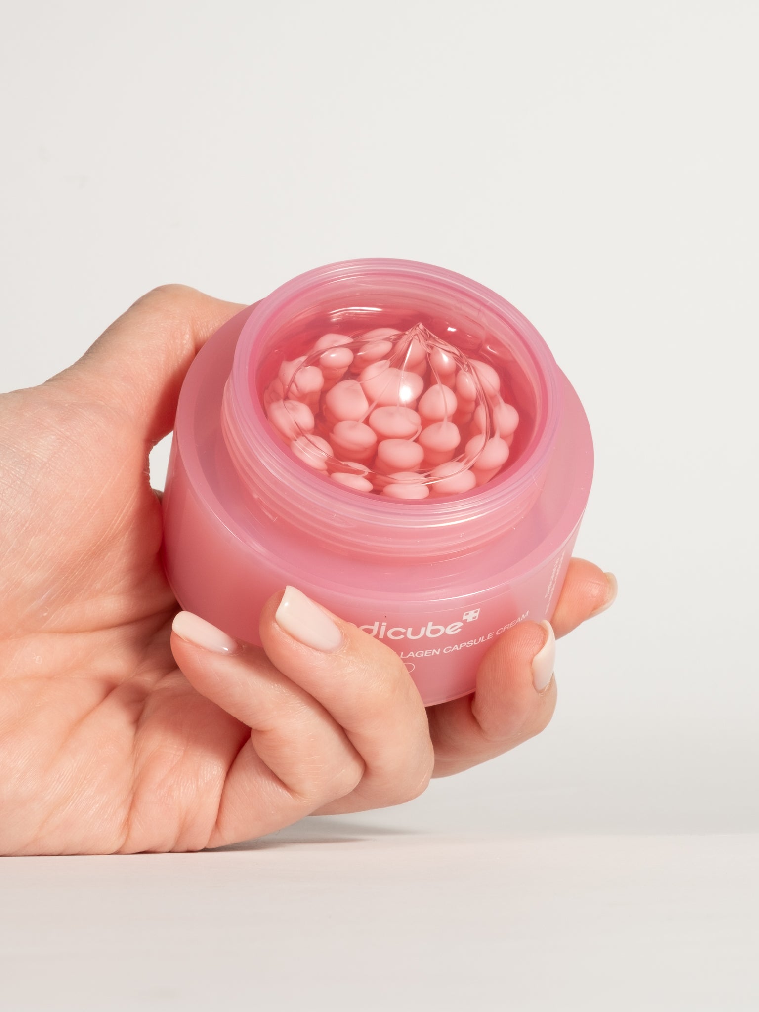 A hand holding an open jar of Medicube PDRN Pink Collagen Capsule Cream, showing the pink capsules suspended in a clear gel.