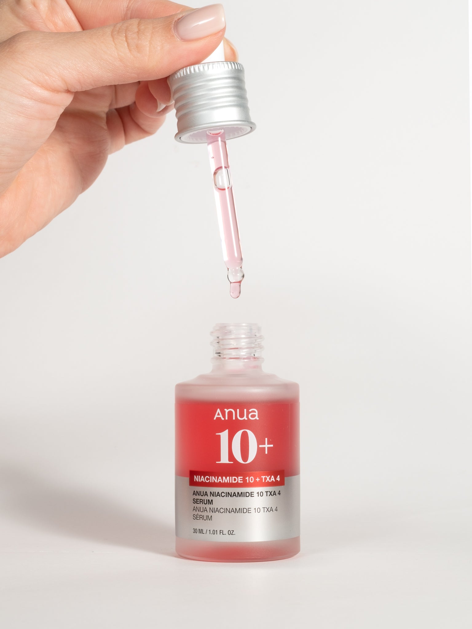 A hand holding a silver dropper with Anua Niacinamide 10 + TXA 4 Serum over an open bottle of the same product.