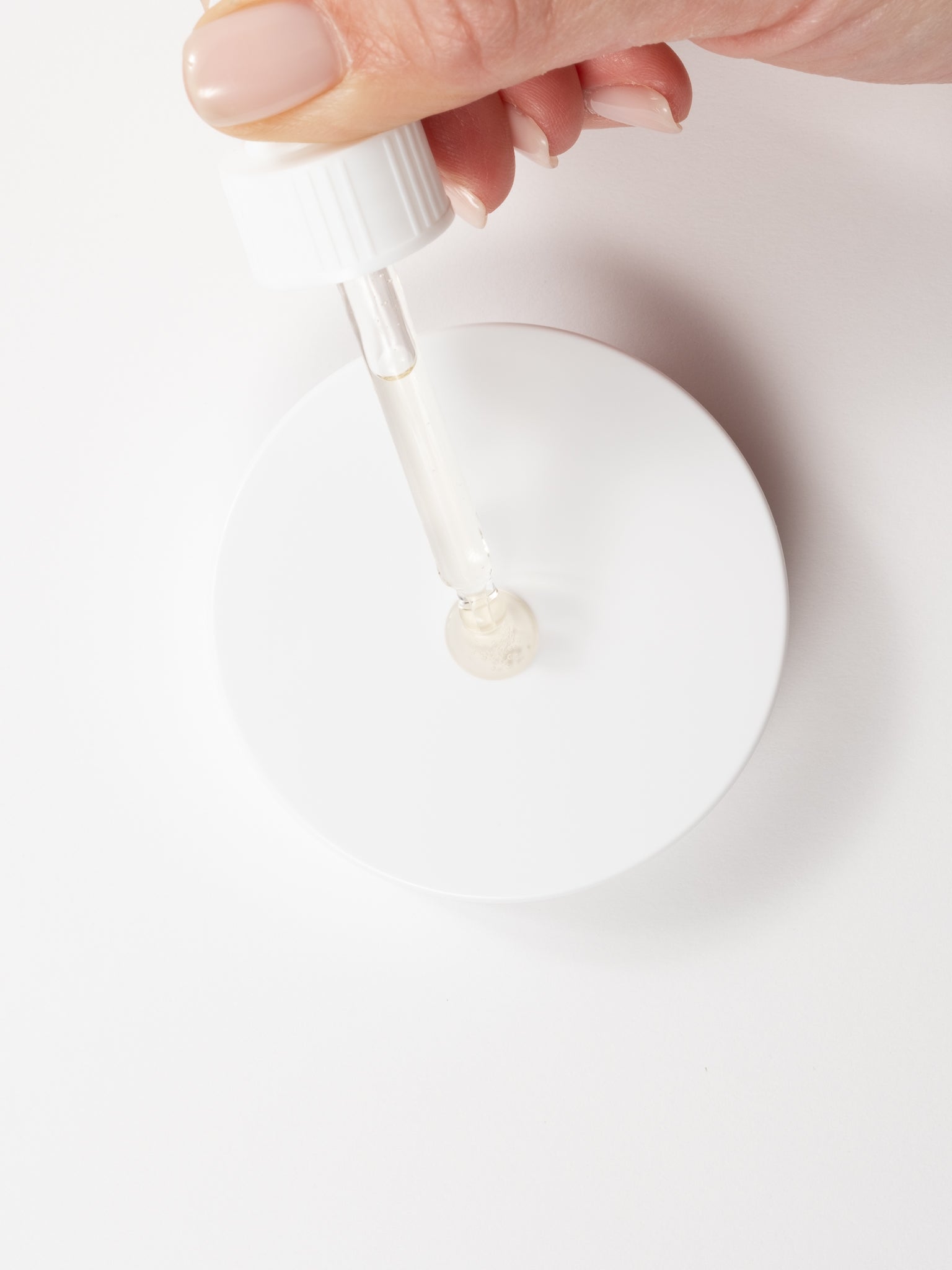 Top-down macro view of a serum droplet being applied from a glass dropper onto a white surface.