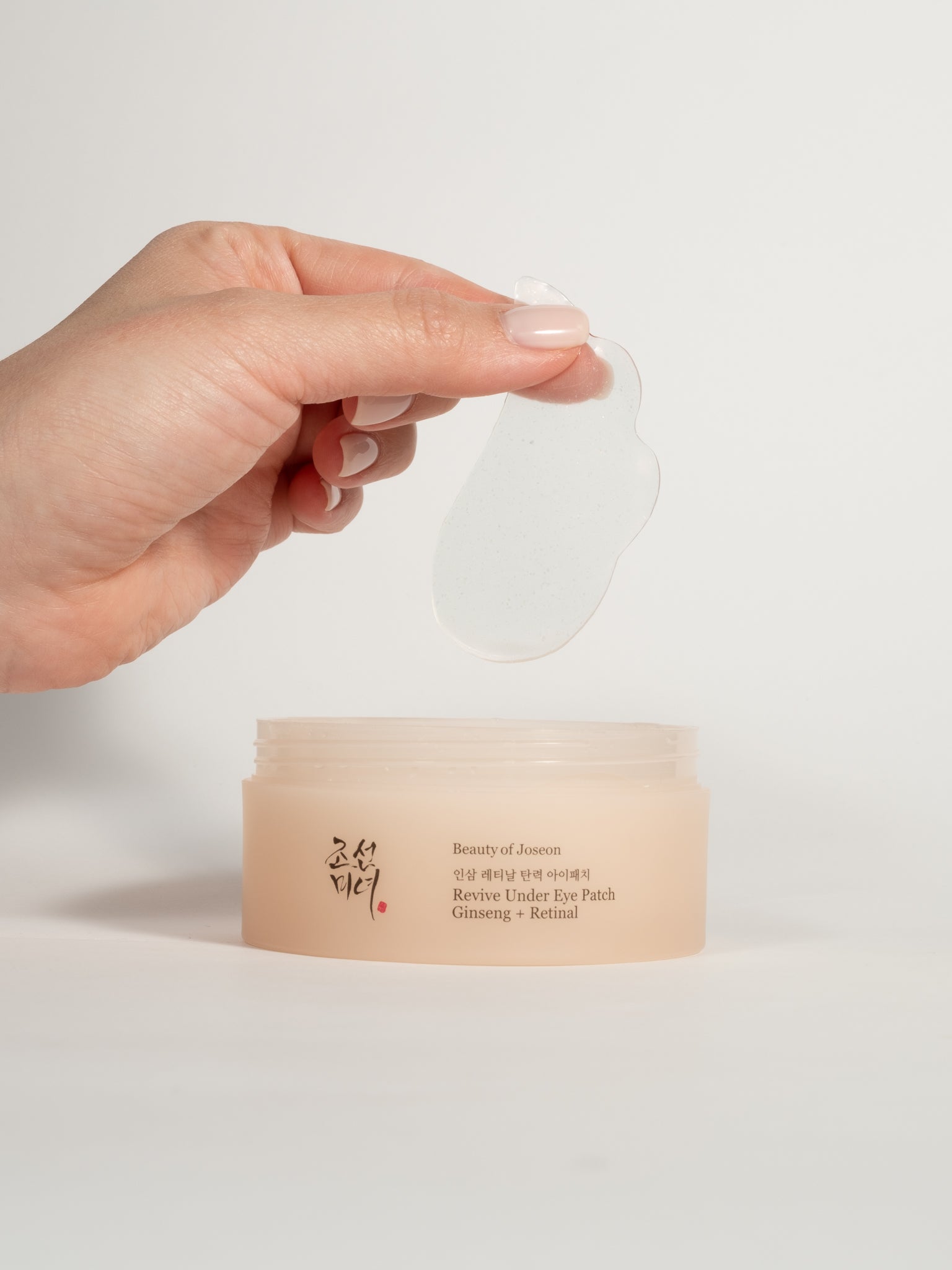 Hand holding a clear eye patch over a jar of Beauty of joseon revive under eye patches on a light background
