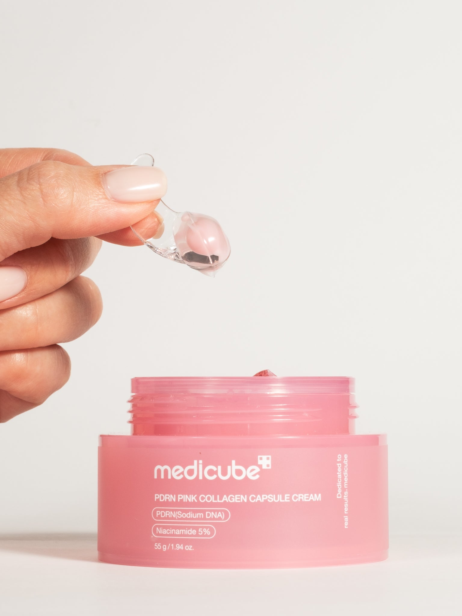A hand using a small clear spatula to scoop a capsule of Medicube PDRN Pink Collagen Capsule Cream from the pink jar.